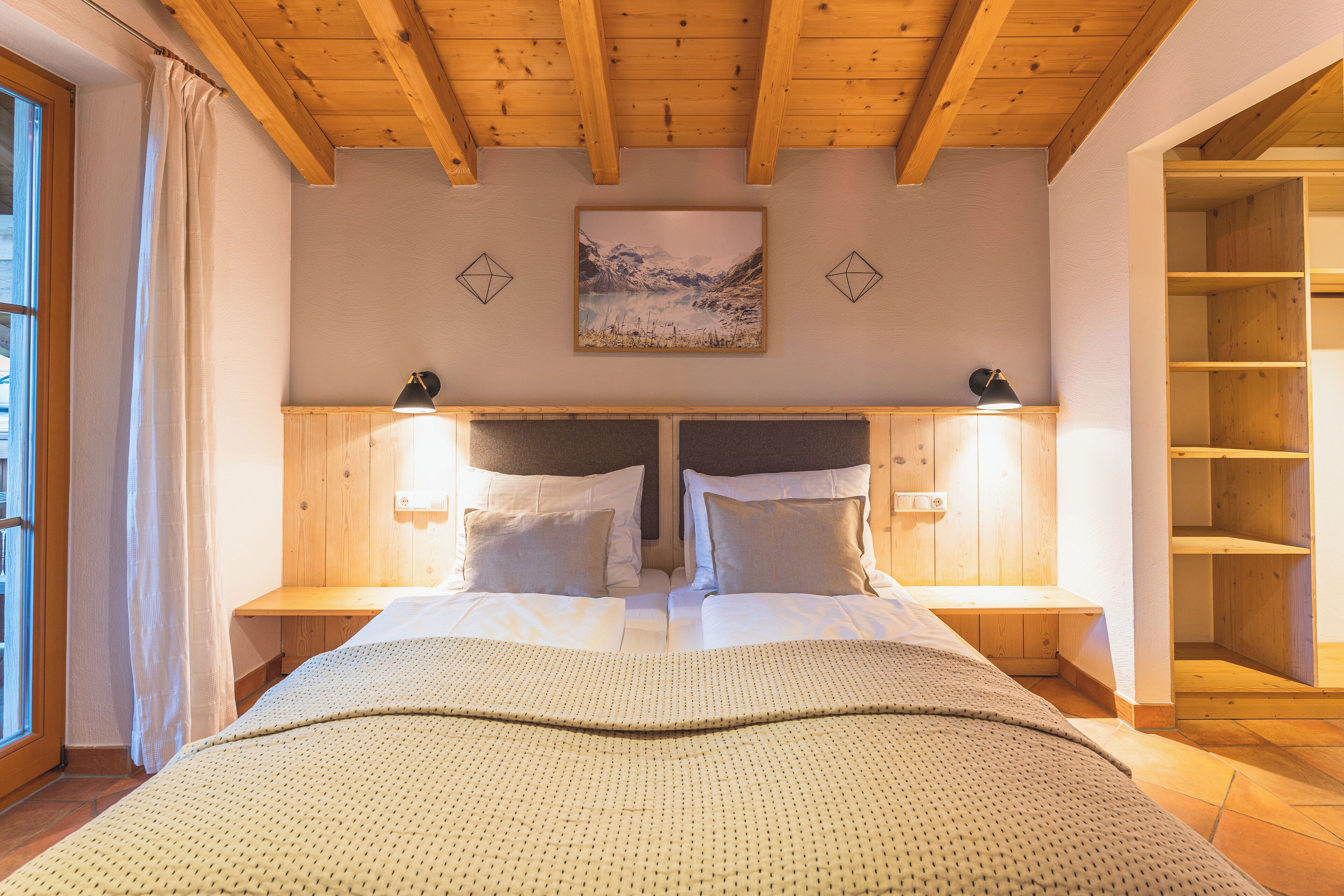 Cama doble con una manta estampada, cabecera de madera, lámparas de noche y un cuadro enmarcado encima.