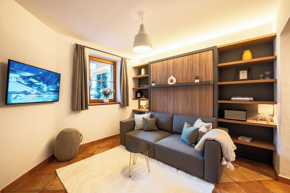 A modern living room with a grey sofa, a white rug, a small side table, a TV mounted on the wall, and a bookshelf with various items.
