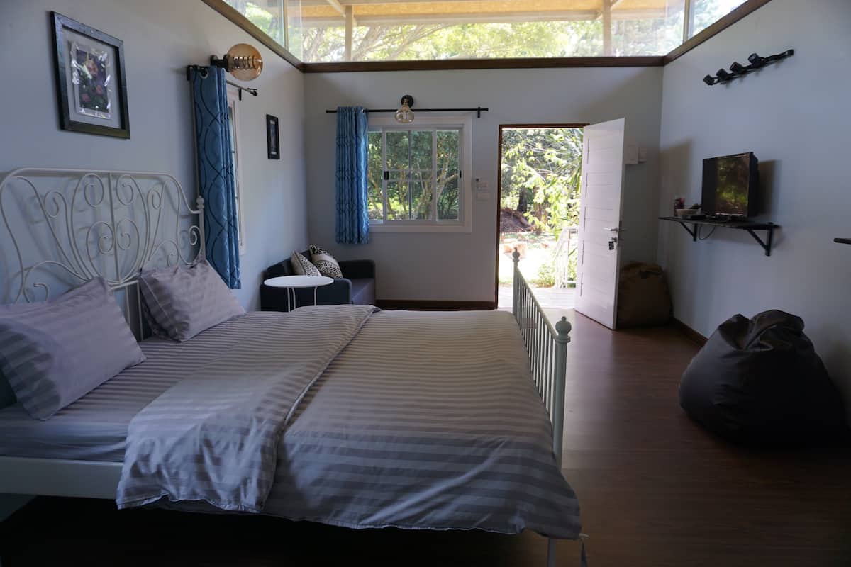 A bedroom with a bed, a small table, a chair, a bean bag, a window with curtains, and a door leading outside.