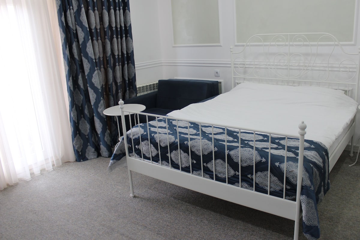 Un letto in metallo bianco con una coperta a letto a motivi blu e bianchi, un tavolino rotondo e una sedia blu scuro in una stanza con pareti bianche e una finestra con tende leggere.