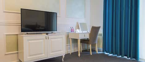 Une chambre d’hôtel avec un téléviseur posé sur un meuble blanc, un bureau avec une chaise et une fenêtre ornée de rideaux bleus.