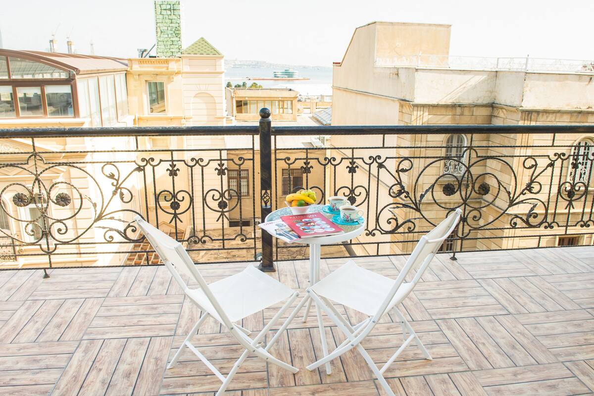 Un balcone con un tavolo apparecchiato per due, con un giornale, una ciotola di frutta e due tazze.