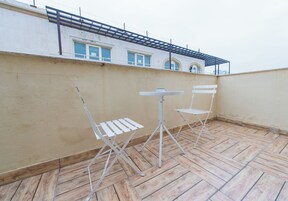 Una terraza en la azotea con piso de madera, dos sillas de metal blanco y una mesita.