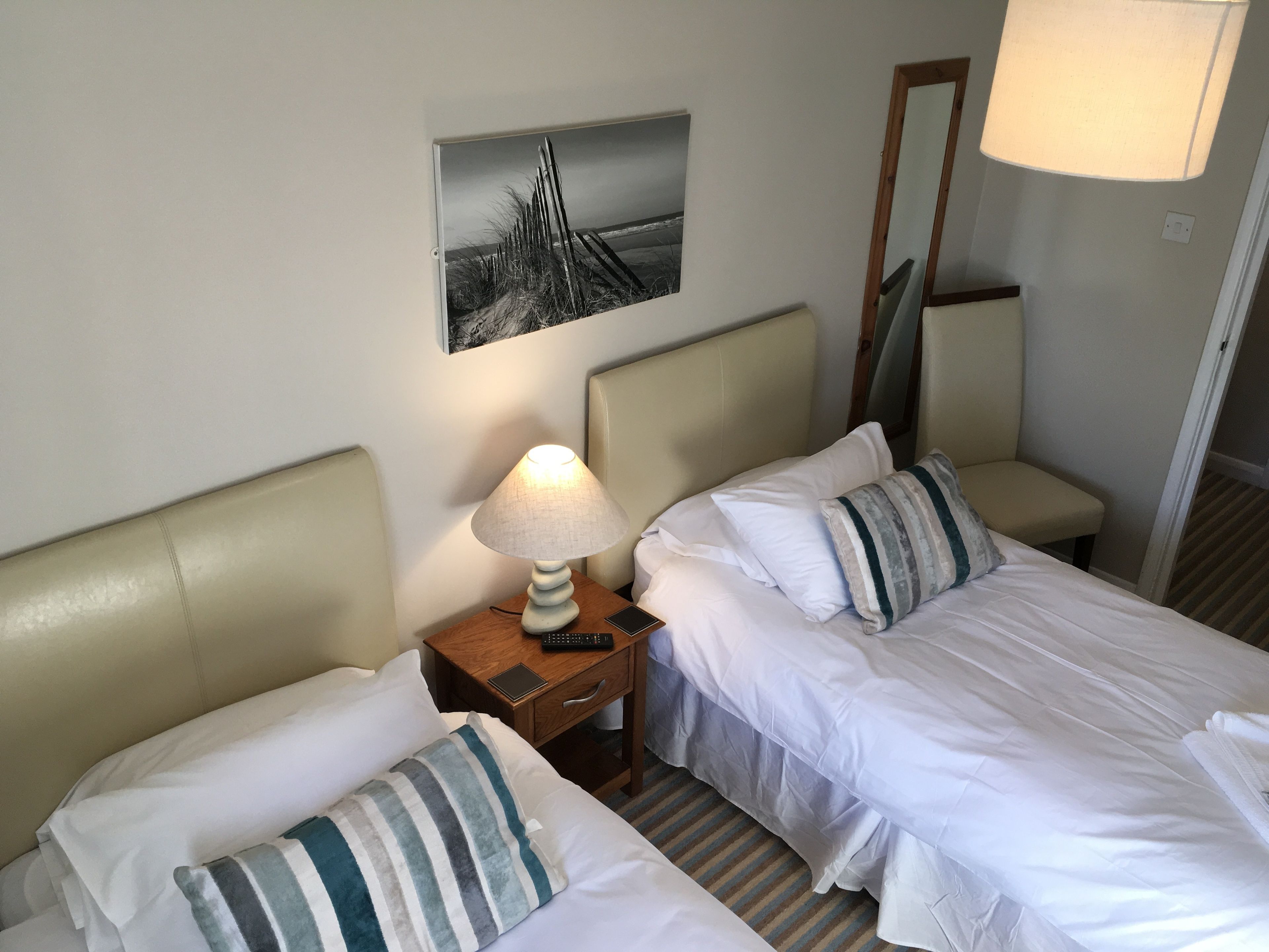 A hotel room with two beds, a nightstand with a lamp, a painting on the wall, and a mirror.