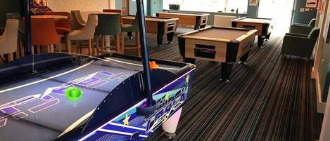 A billiards table with a starry design, a pool table, and a digital scoreboard in a room with striped carpet and pendant lighting.