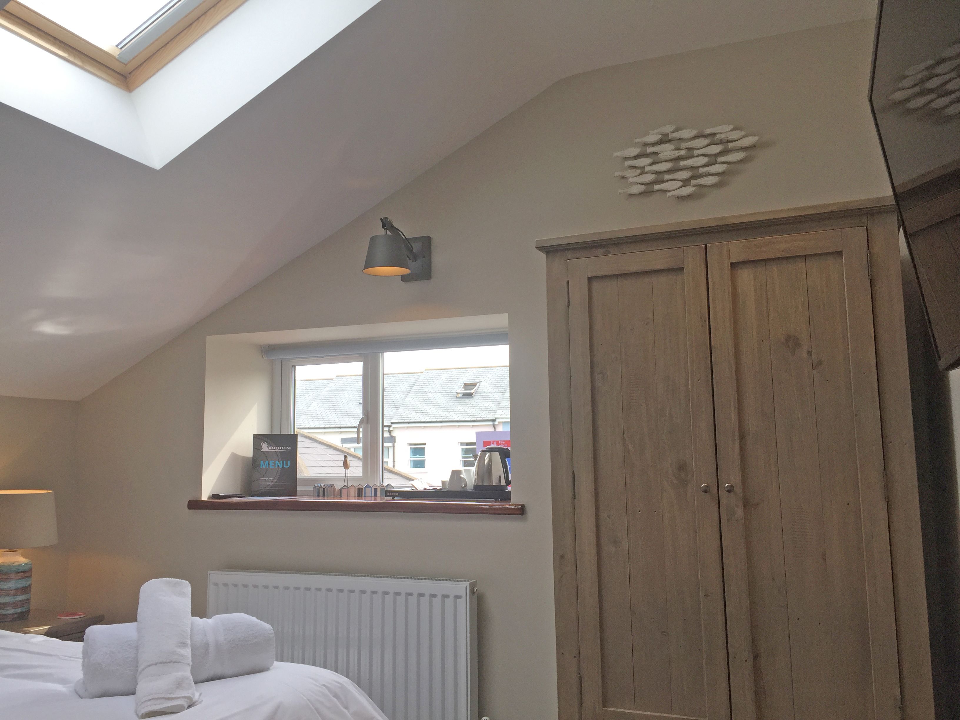 A bedroom with a skylight, a wooden wardrobe, a radiator, and a window with a view of buildings outside.