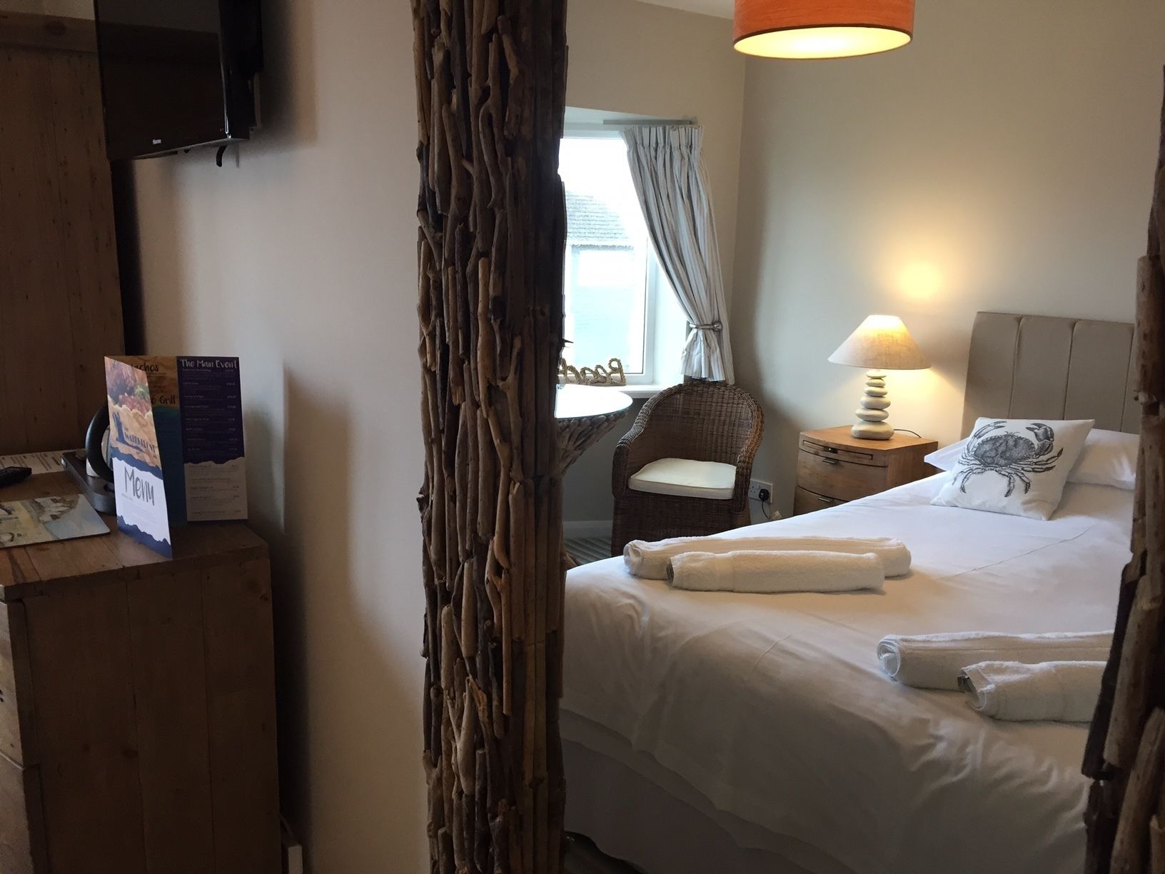 A hotel room with a bed, a chair, a nightstand with a lamp, a wooden wall feature, and a window with curtains.
