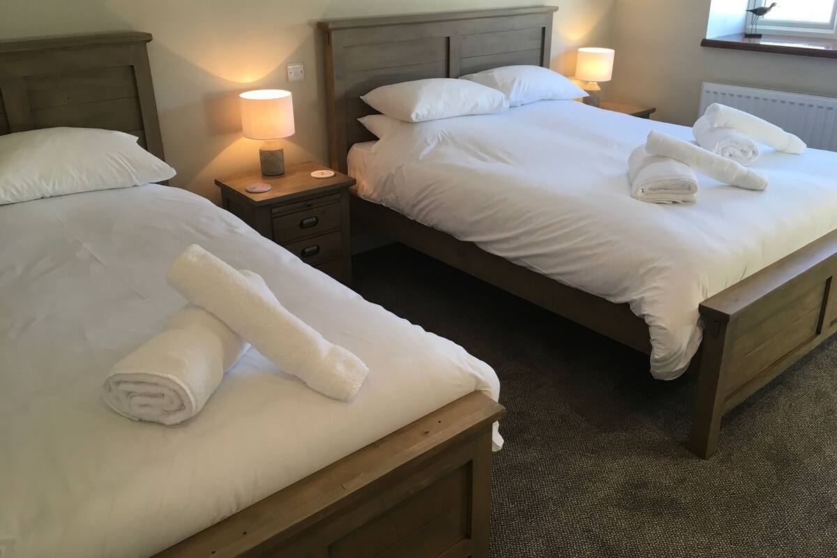 Quarto de hotel com duas camas, cada uma com roupa de cama e toalhas brancas. Há mesas de cabeceira com abajures e uma janela com vista.