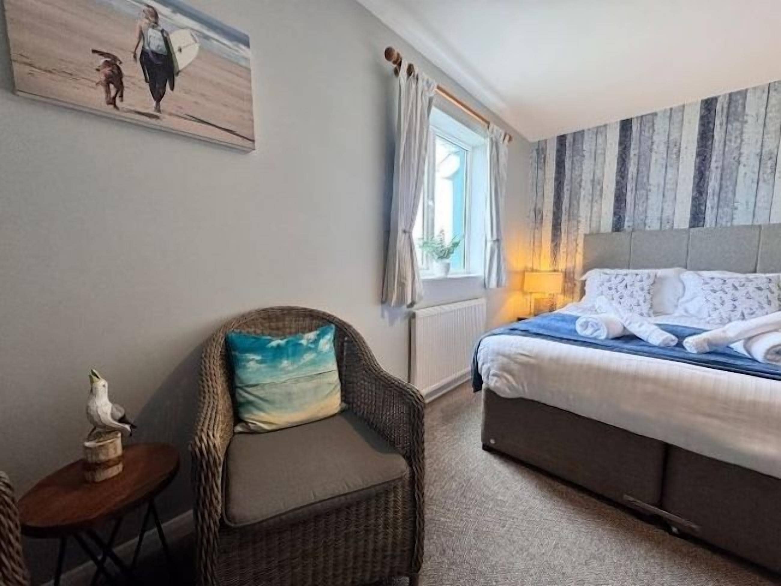 Ein Schlafzimmer mit einem Bett, einem Sessel mit Kissen, einem kleinen Tisch und einem Fenster mit Vorhängen.
