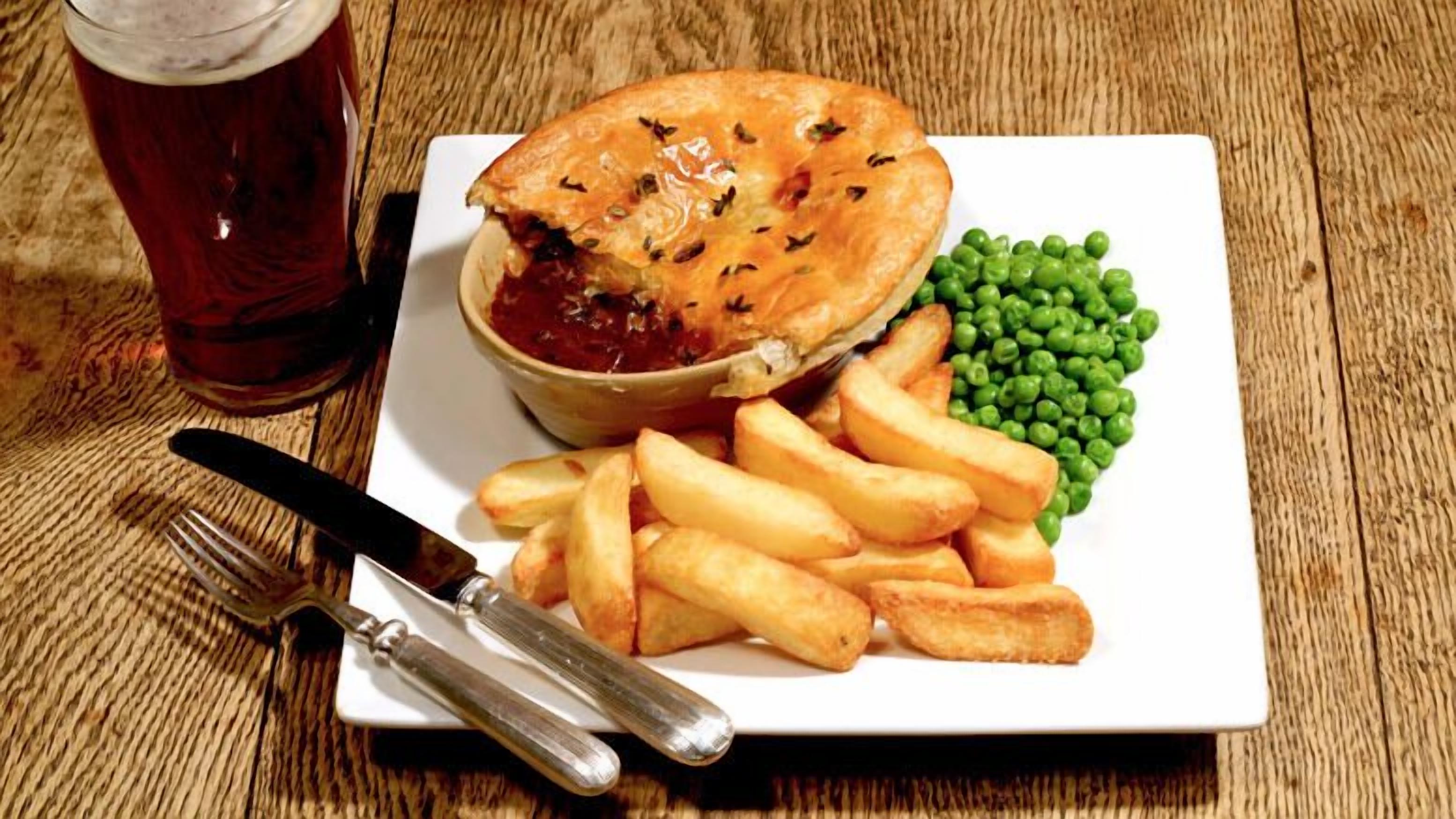 A meal consisting of a pie, peas, and fries on a white plate, accompanied by a drink and cutlery on a wooden table.