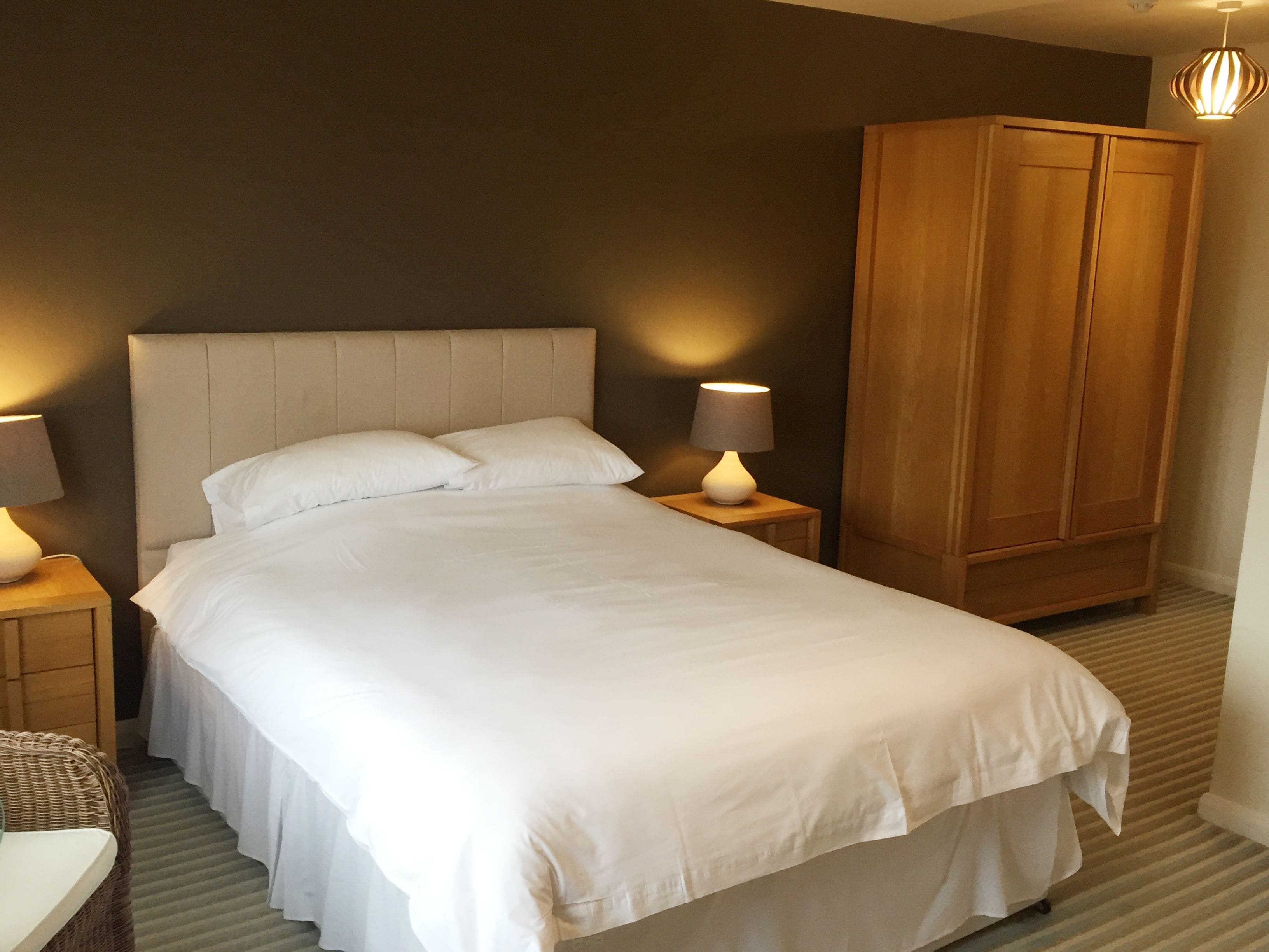 A neatly made bed with white linens, two bedside lamps, a wooden wardrobe, and a wicker chair.