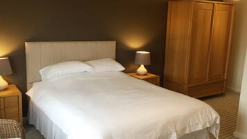 A neatly made bed with white linens, two bedside lamps, a wooden wardrobe, and a wicker chair.
