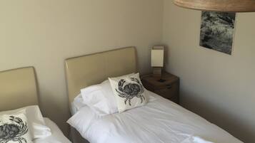 Une chambre d’hôtel avec deux lits simples, chacun recouvert d’une courtepointe blanche et agrémenté d’un oreiller orné d’un motif de crabe. Une table de chevet avec une lampe se trouve entre les lits, et un tableau encadré est accroché au mur.