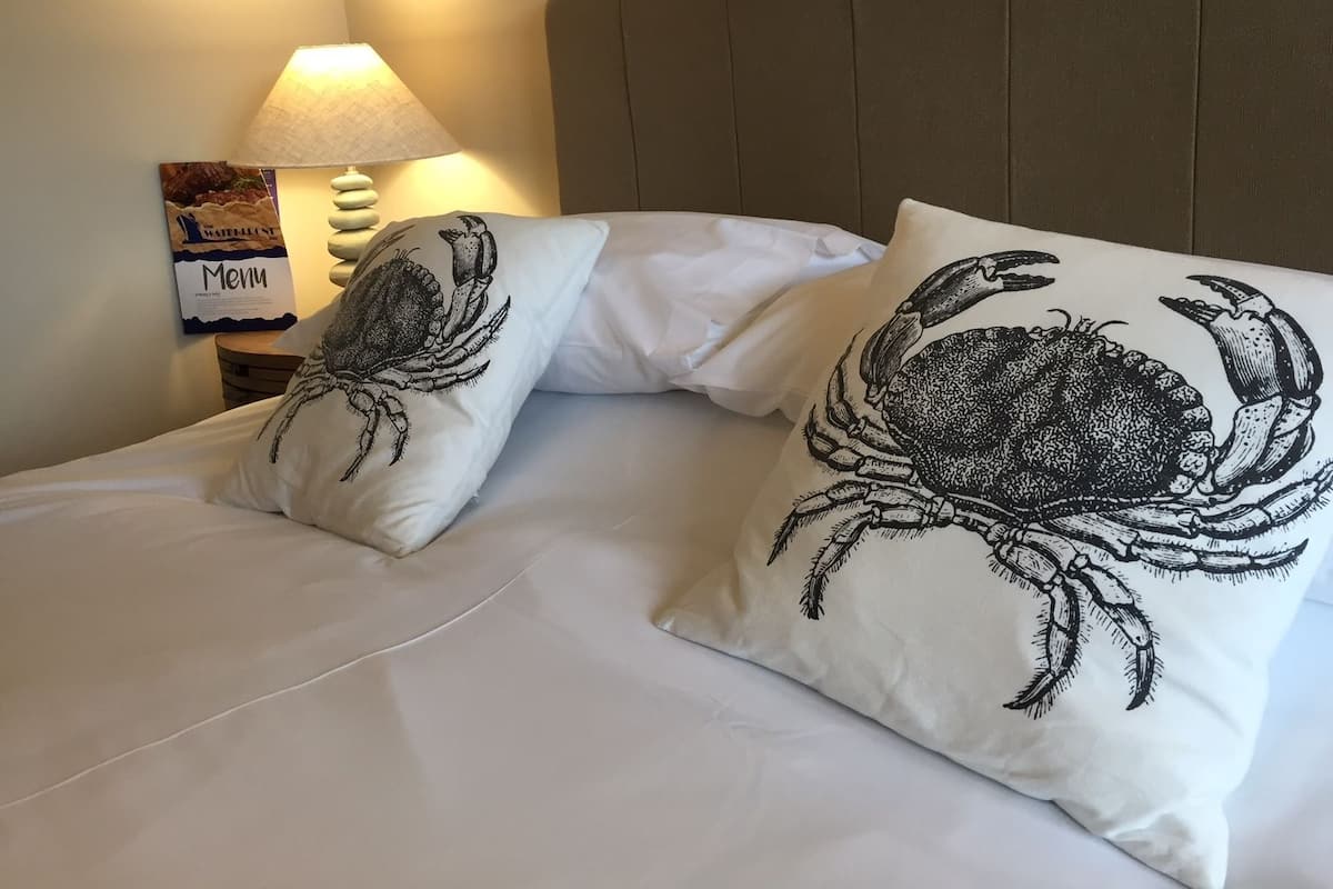 Uma cama com roupa de cama branca e duas almofadas com o desenho de caranguejos. Uma luminária e um letreiro de menu estão visíveis na parede ao lado da cama.