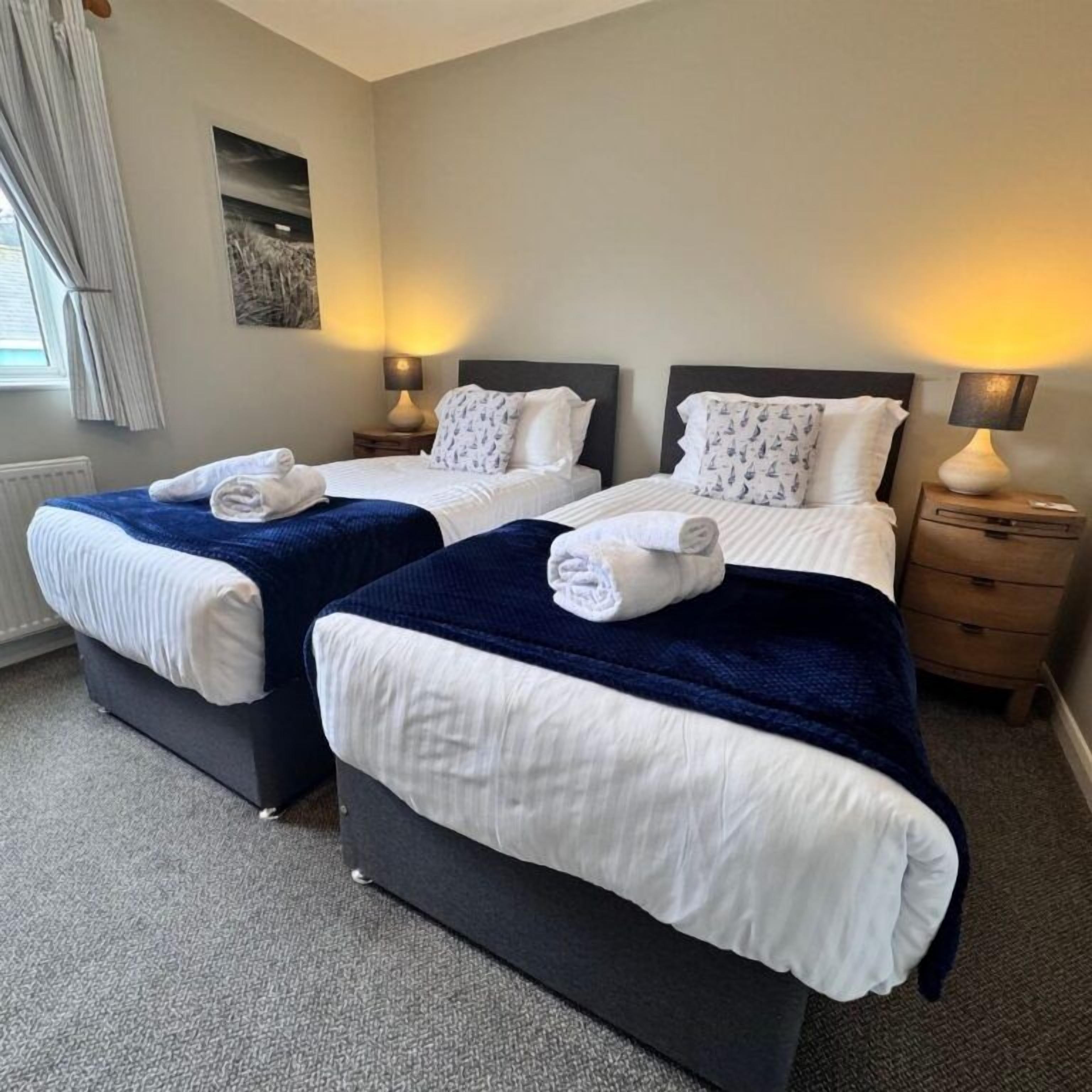 Habitación de hotel con dos camas, cada una con sábanas blancas y una manta azul oscuro, una mesita de noche con lámpara y una ventana con cortinas.
