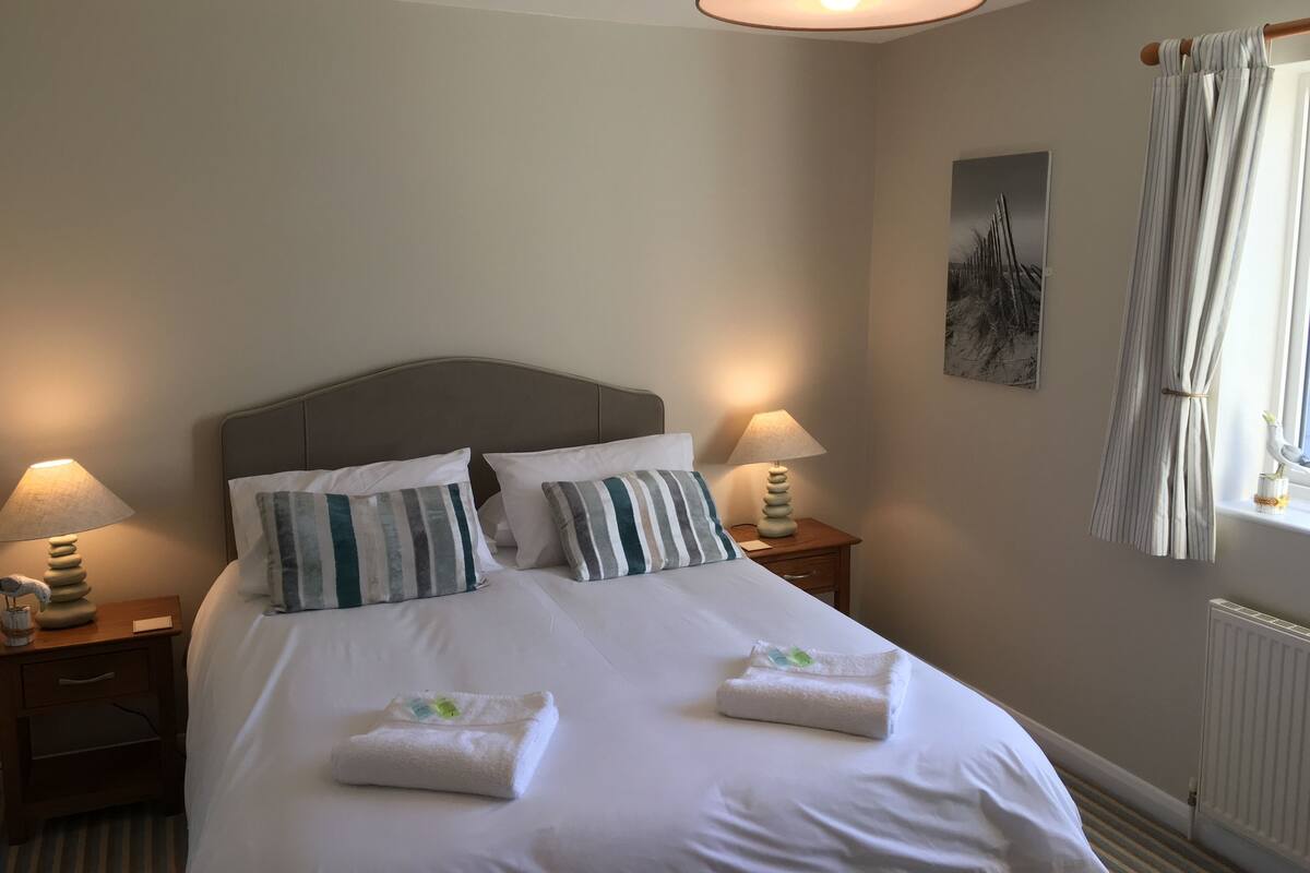 Cama bem arrumada com duas almofadas, abajur e mesa pequena com um objeto decorativo.