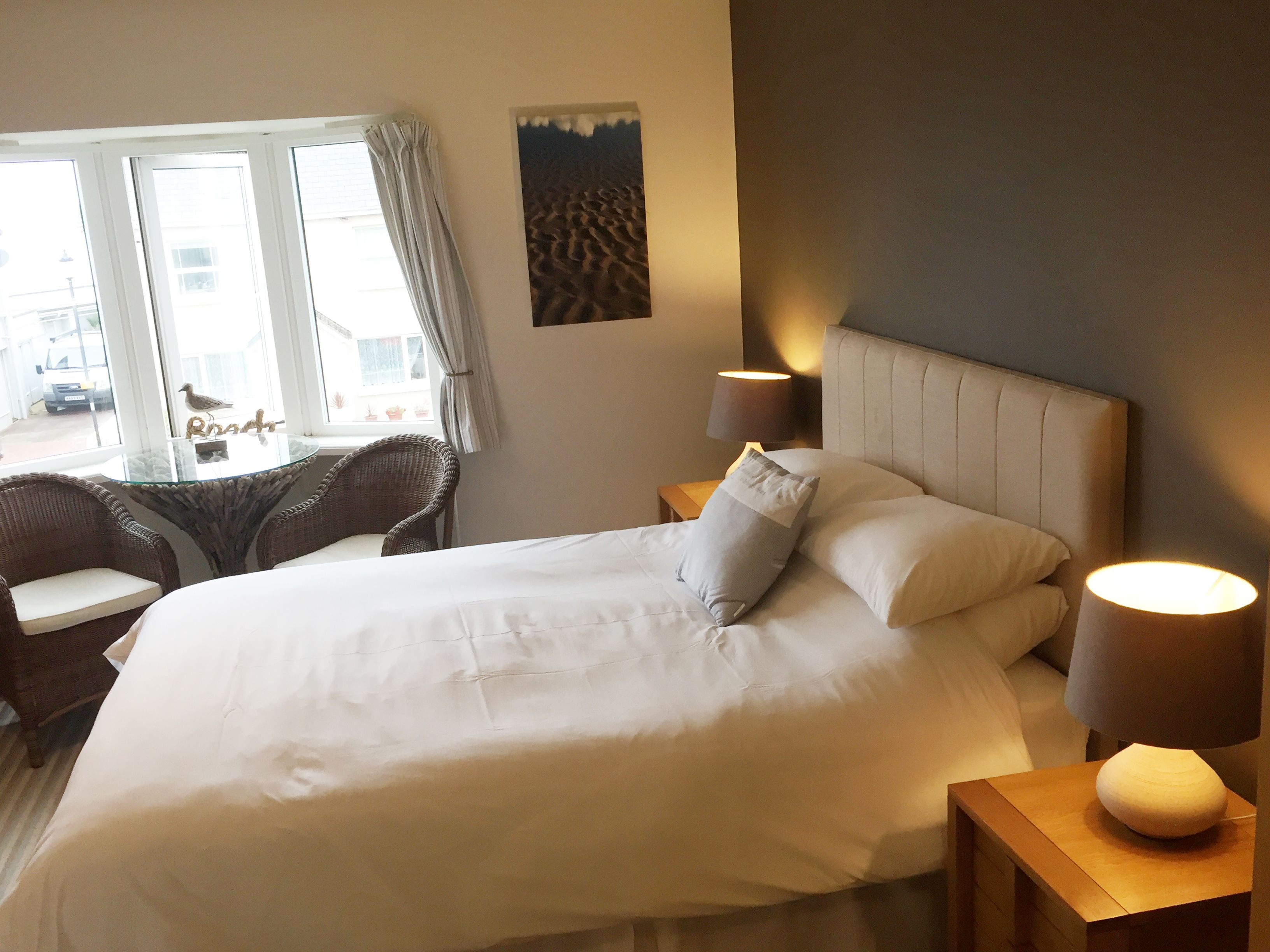 A neatly made bed with pillows, a bedside lamp, a window with curtains, and a wicker chair.