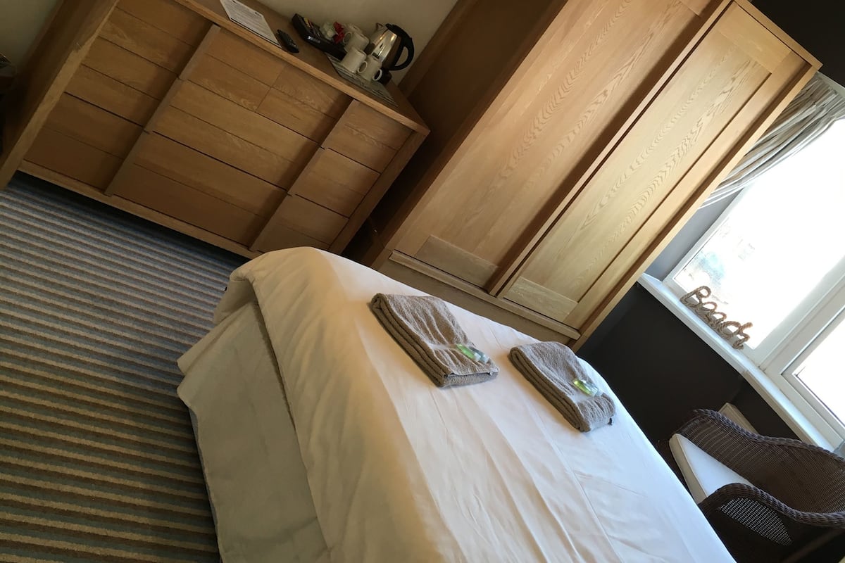 Quarto de hotel com cama, guarda-roupa de madeira, escrivaninha com abajur e um quadro na parede.