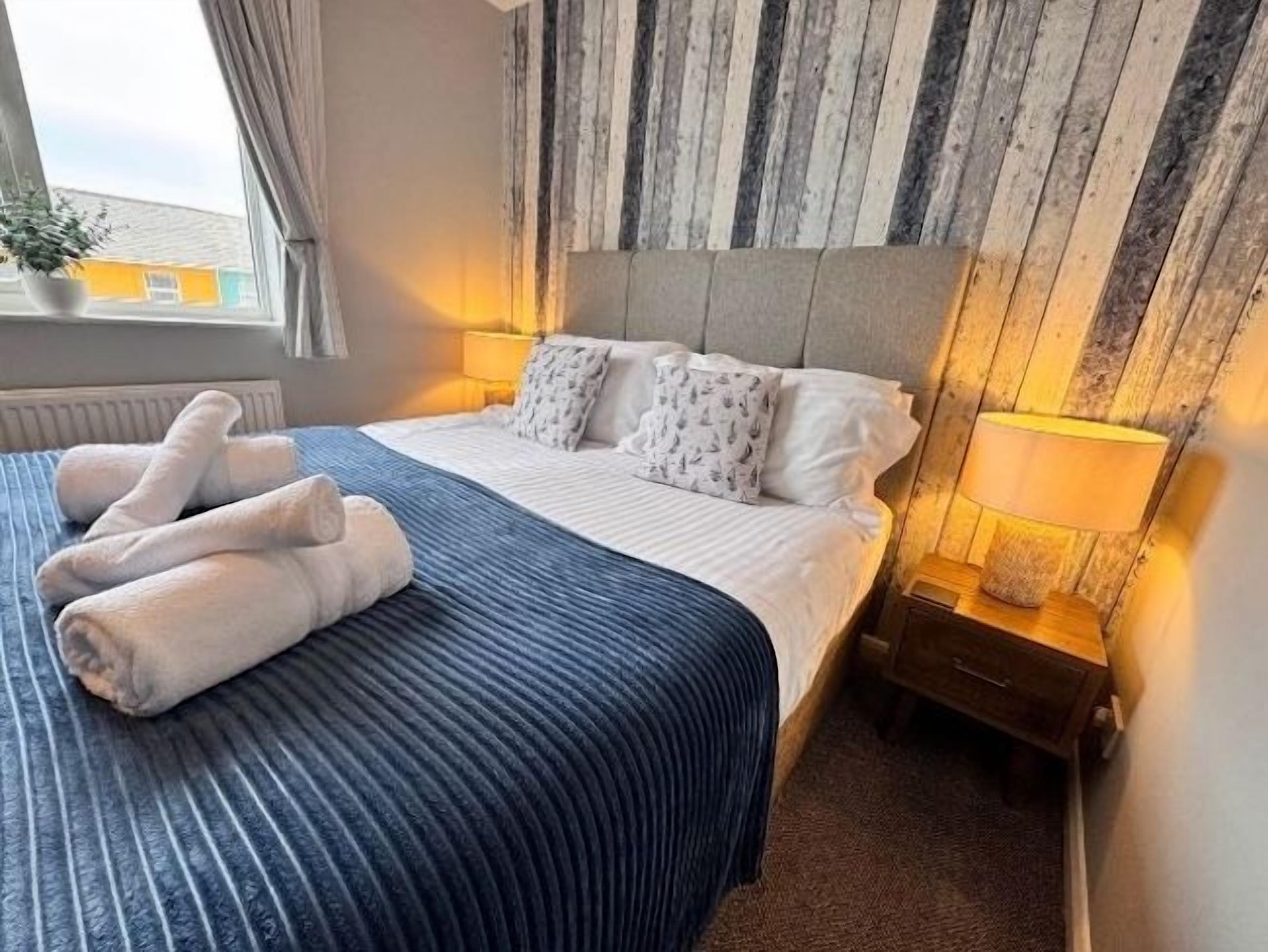 Una habitación de hotel con cama, mesita de noche, lámpara y ventana con cortinas.