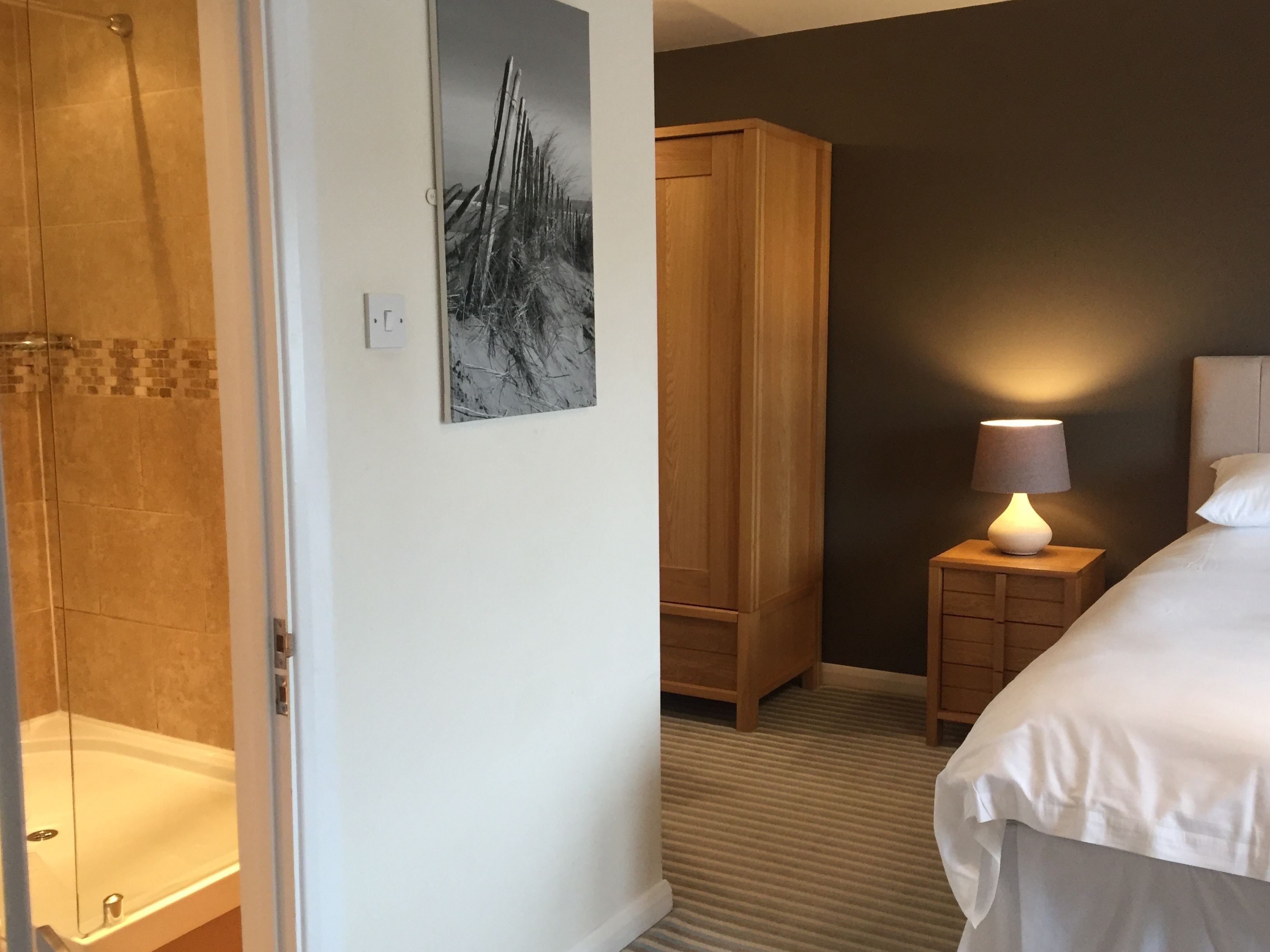 Ein Hotelzimmer mit Dusche, einem Bett mit weißer Bettwäsche, einem Nachttisch mit Lampe, einem Holzschrank und einem an der Wand hängenden Bild.