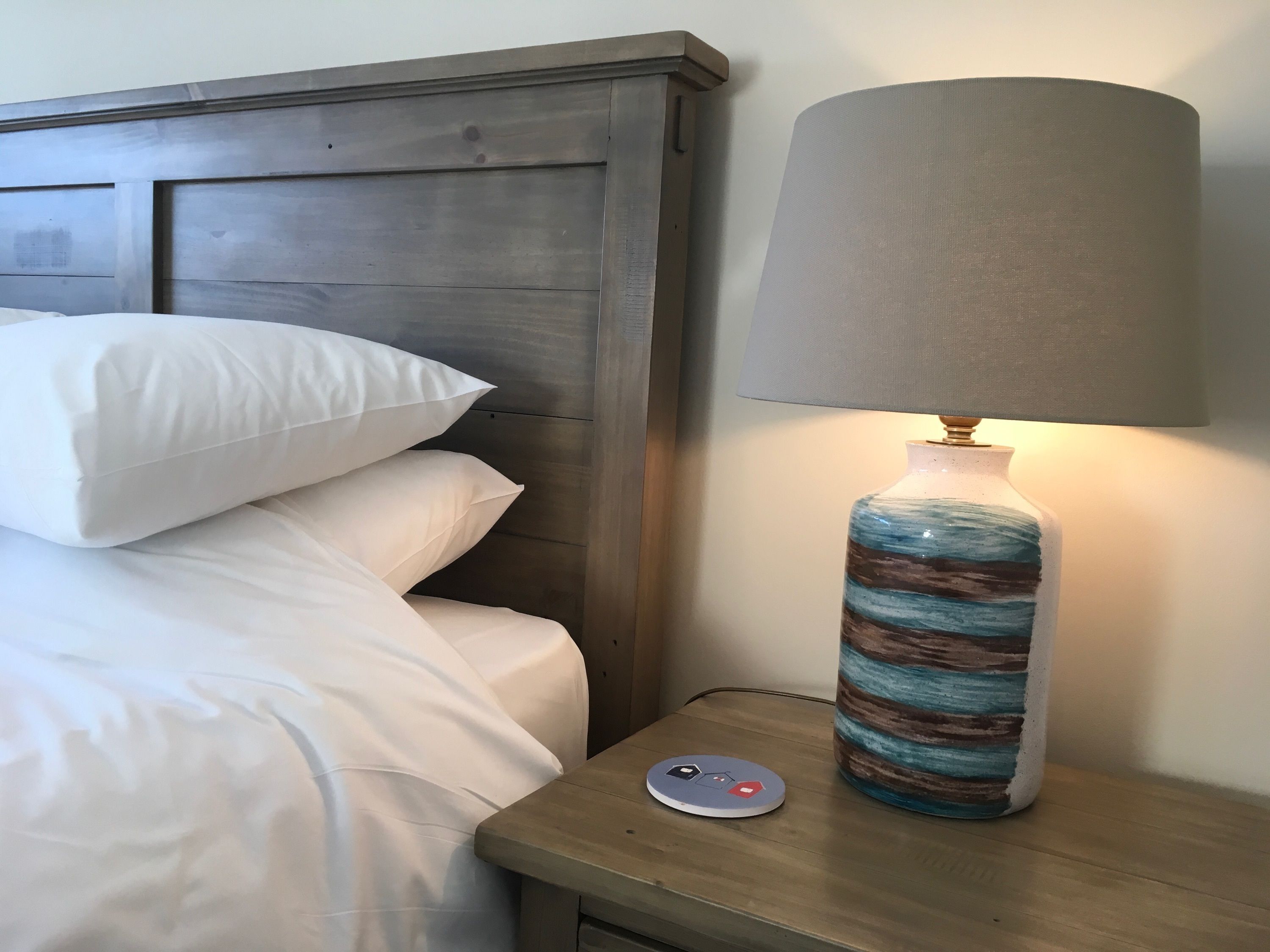 A neatly made bed with white linens and pillows, a wooden nightstand with a lamp and a power outlet, and a wall with a wooden headboard.