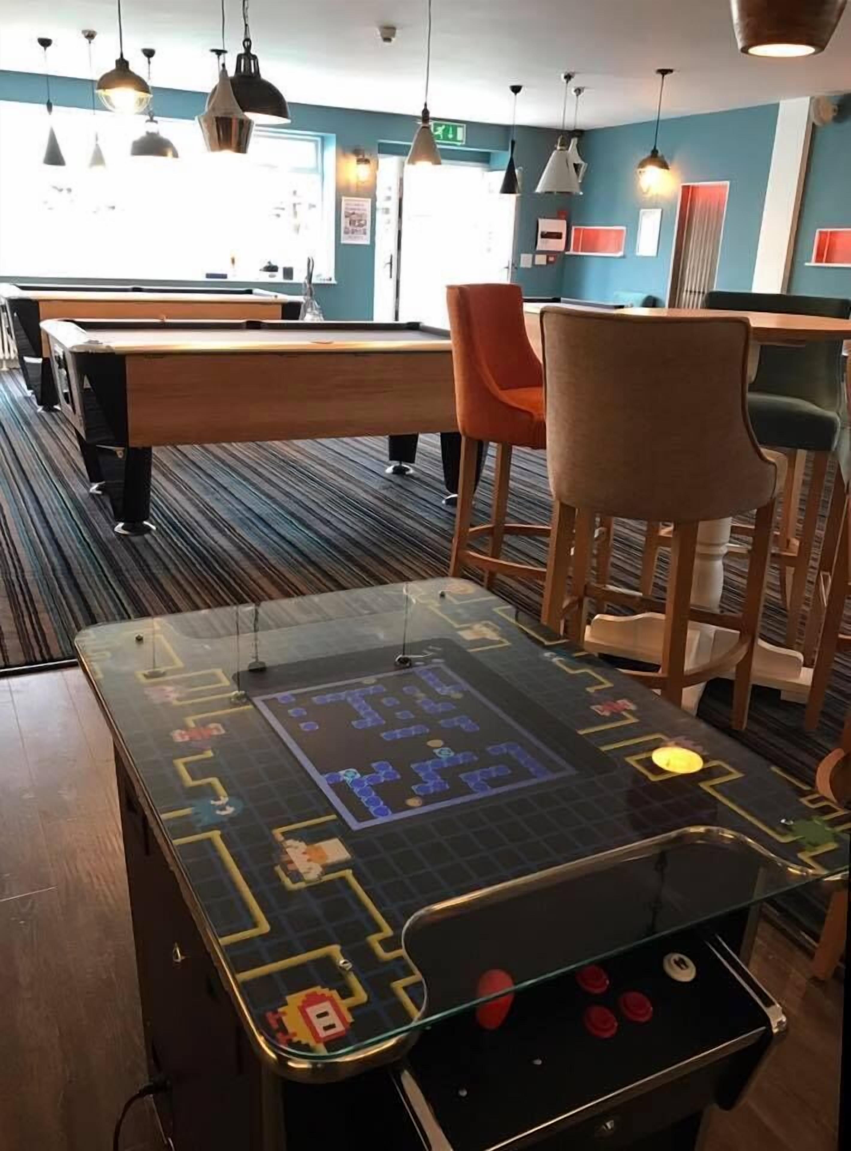 Una mesa de billar con superficie de vidrio y una máquina de pinball en una habitación con pared azul, muebles de madera e iluminación colgante.