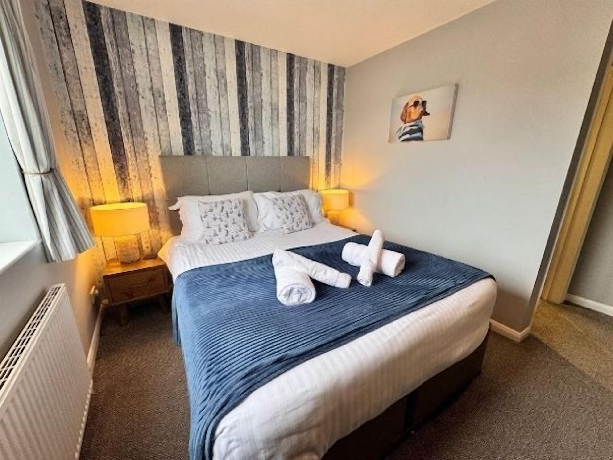Una habitación de hotel con una cama grande, un cabecero con estampado a rayas, una mesita de noche con lámpara y un cuadro enmarcado en la pared.