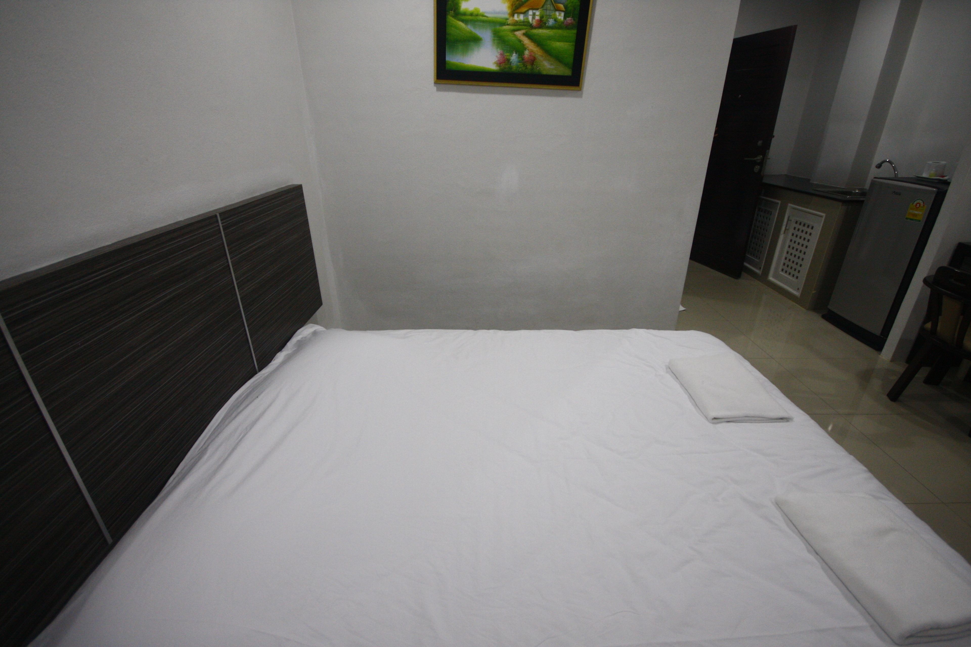 A neatly made bed in a hotel room with a headboard, a painting on the wall, and a kitchenette in the background.