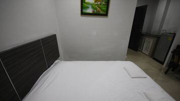 A neatly made bed in a hotel room with a headboard, a painting on the wall, and a kitchenette in the background.