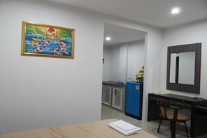 A hotel room with a bed, a desk with a chair, a large mirror, a painting on the wall, and a small kitchenette area.