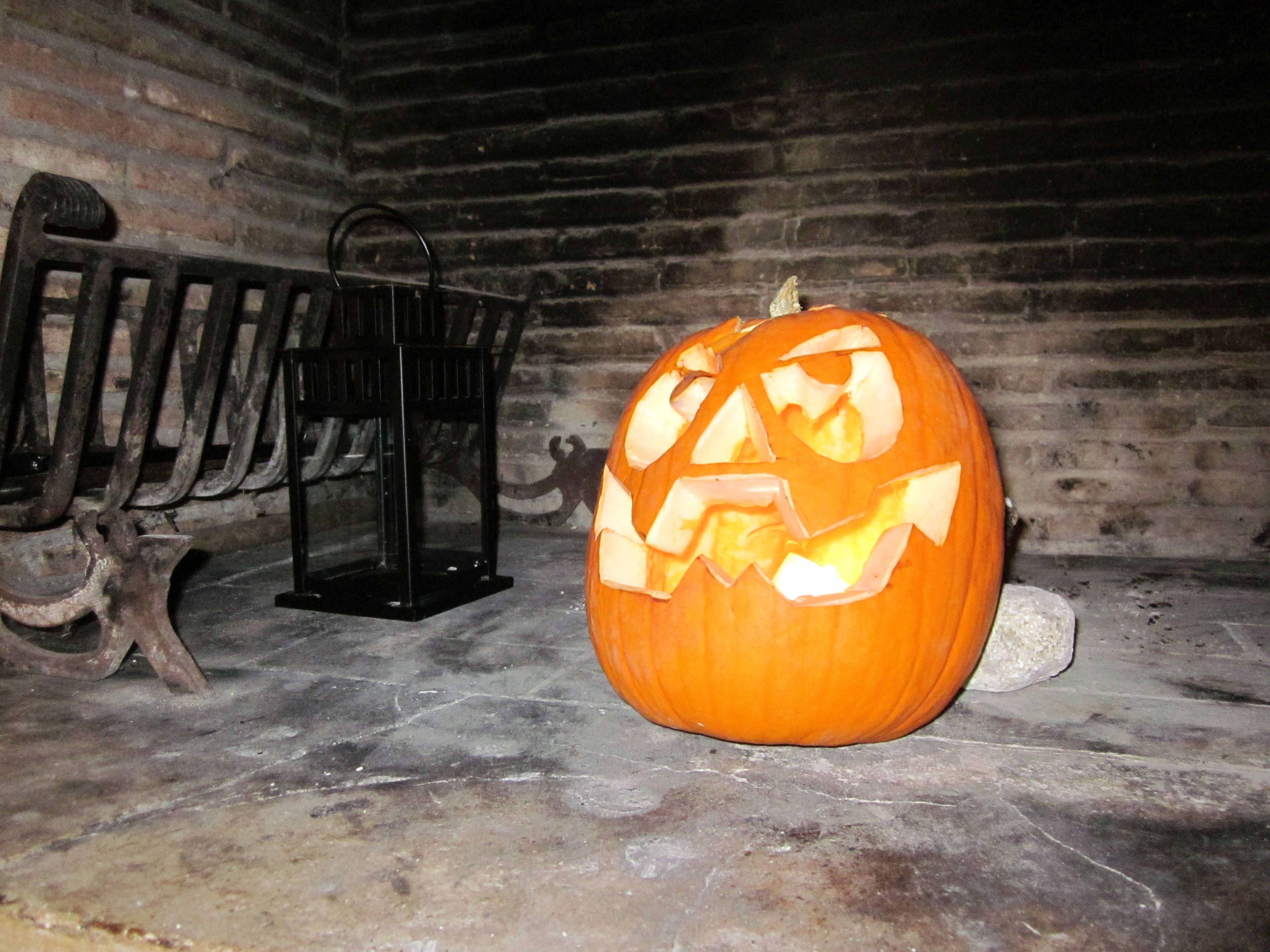 A carved pumpkin with a menacing face, placed on a stone floor next to a metal lantern in a rustic room with wooden walls.