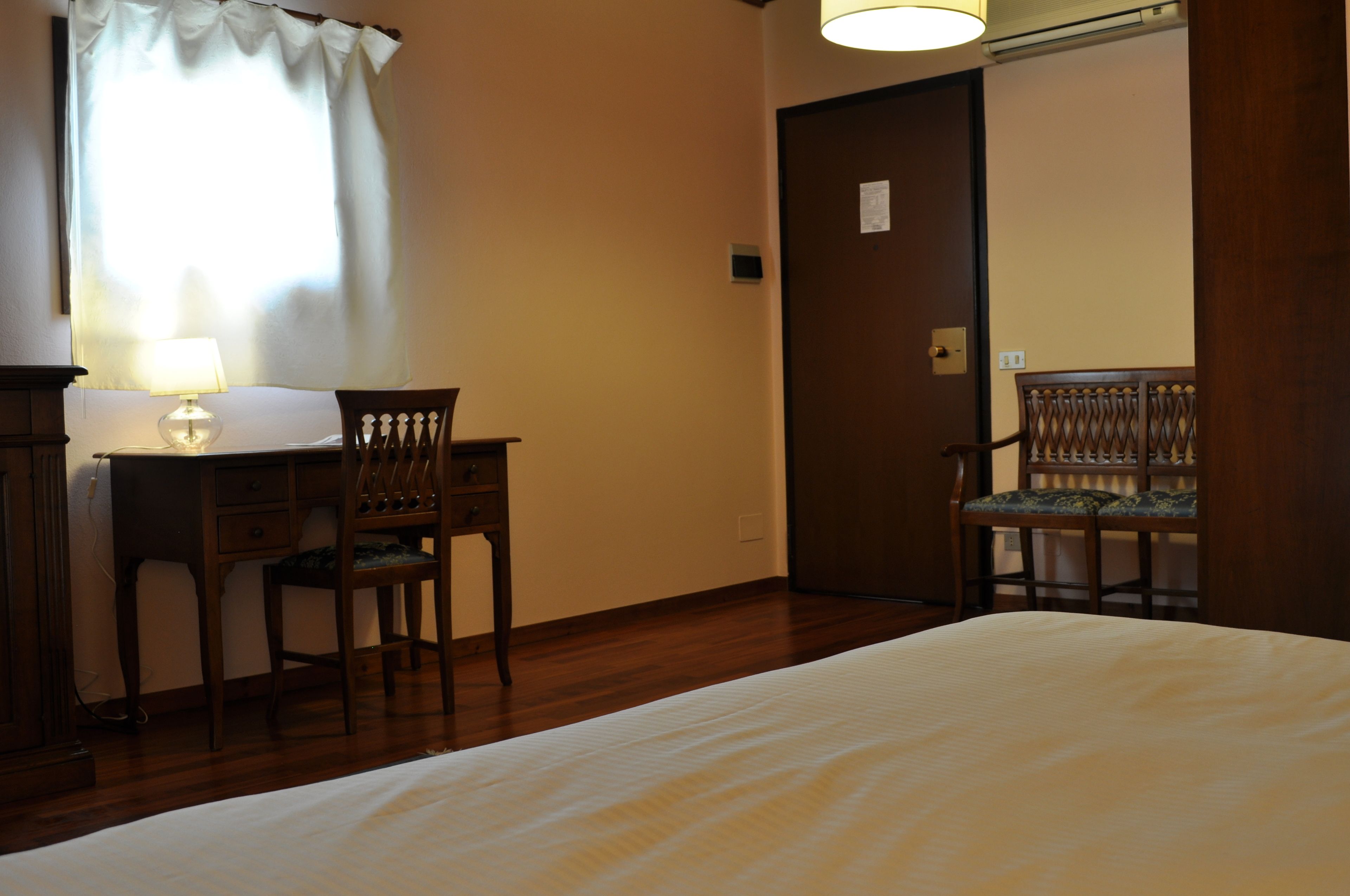 A hotel room with a bed, a desk with a lamp, two chairs, and a door.
