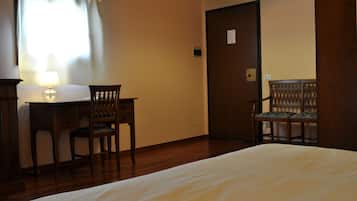 A hotel room with a bed, a desk with a lamp, two chairs, and a door.