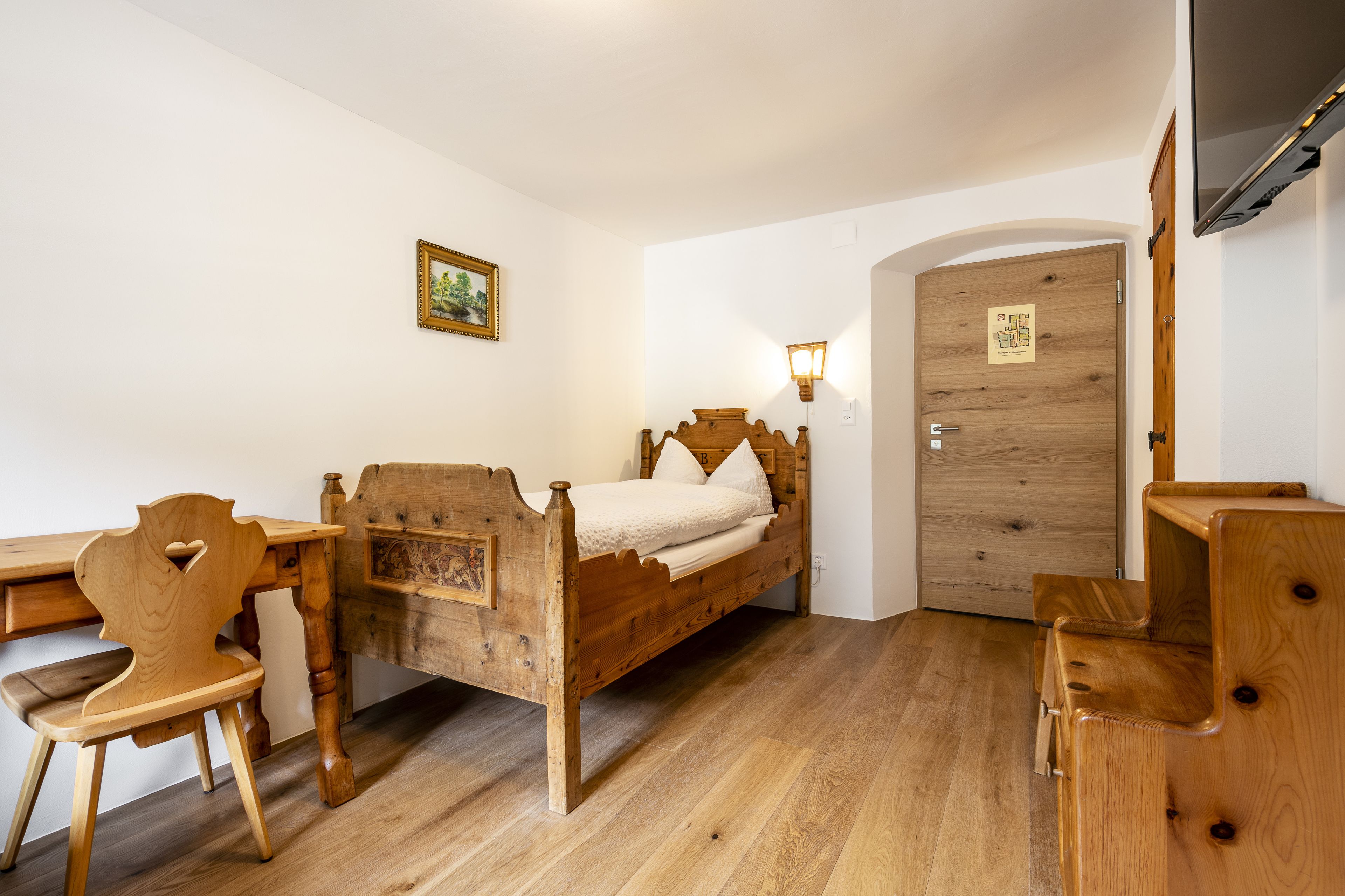 Una cama de madera con cabecera, un escritorio de madera y una silla en una habitación con paredes blancas y suelo de madera.