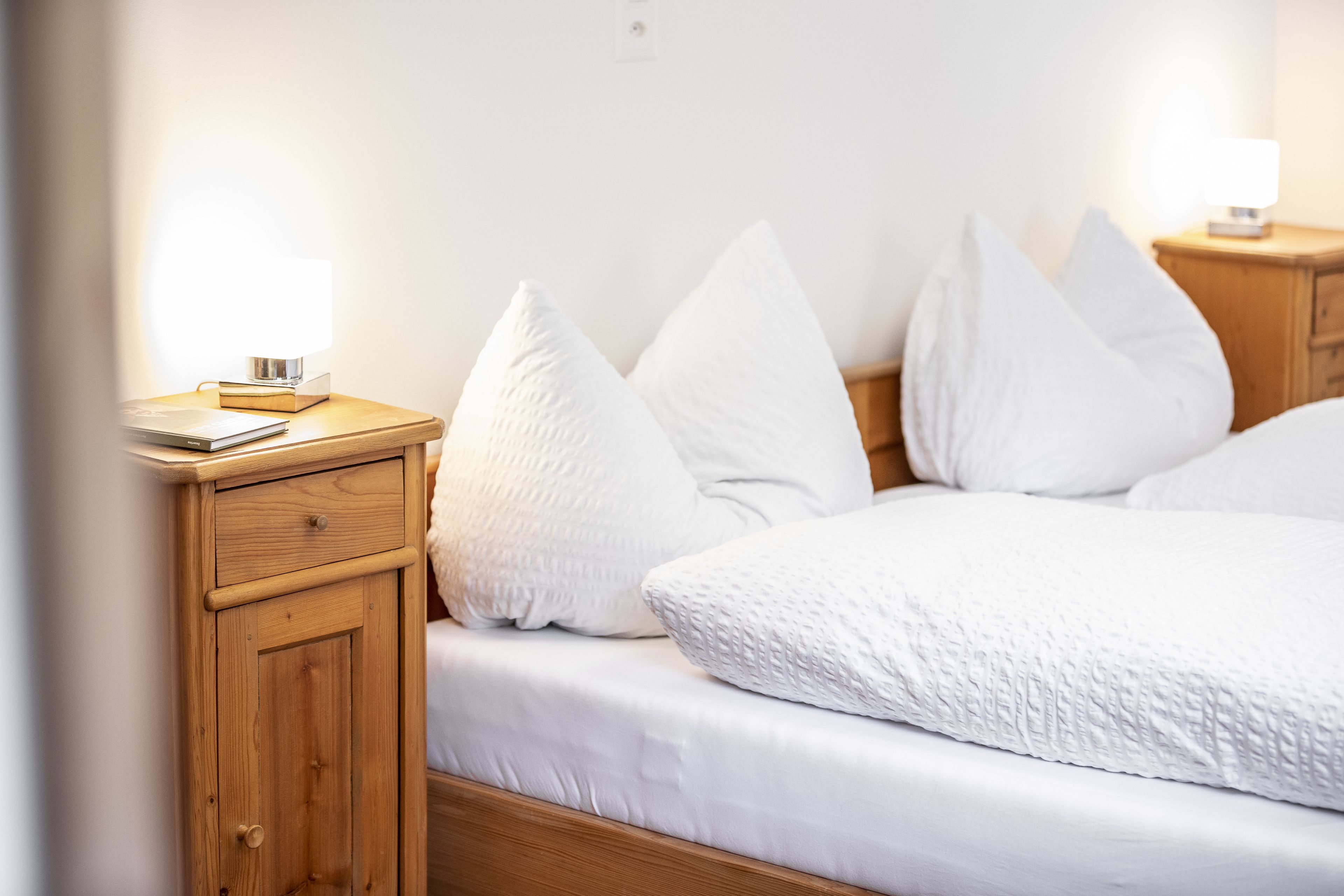 A neatly made bed with white bedding and pillows, a wooden nightstand with a lamp, and a small drawer unit.