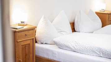 A neatly made bed with white bedding and pillows, a wooden nightstand with a lamp, and a small drawer unit.