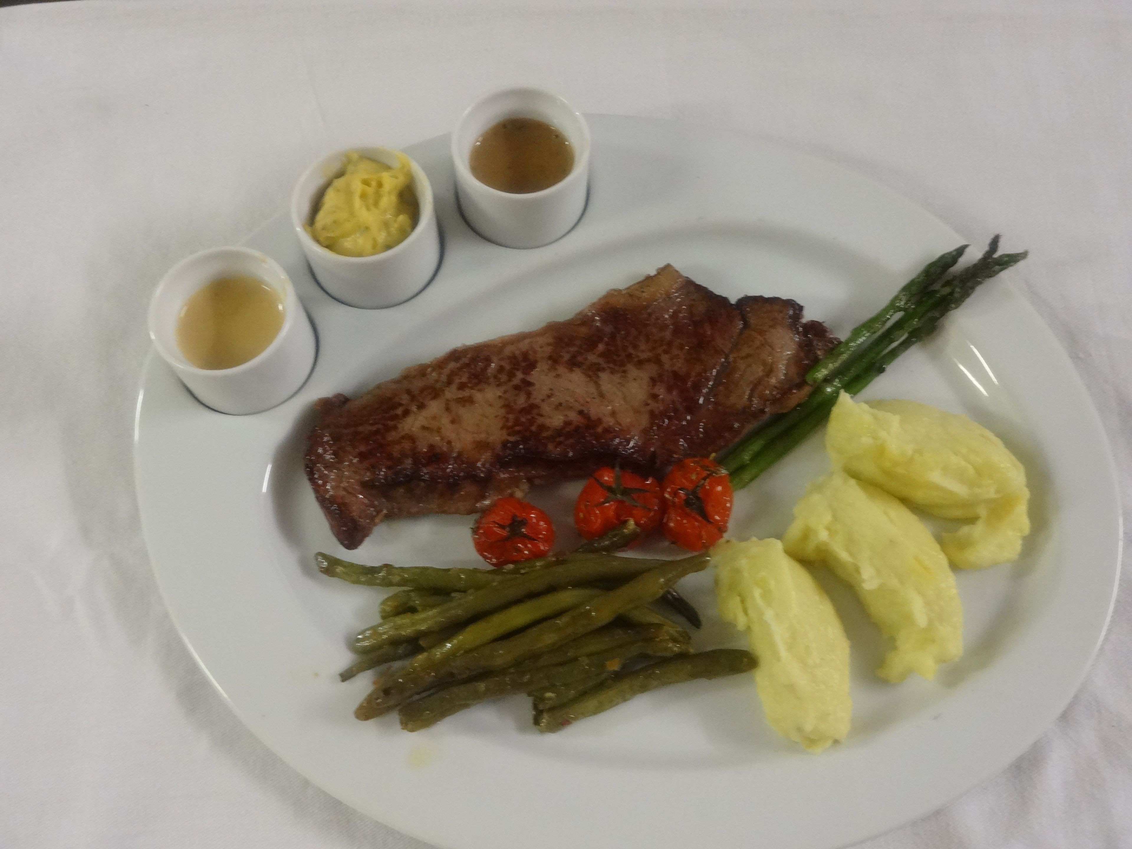 Ein Teller mit einem Steak, Spargel, Cocktailtomaten, Kartoffelpüree und drei kleinen Soßen.