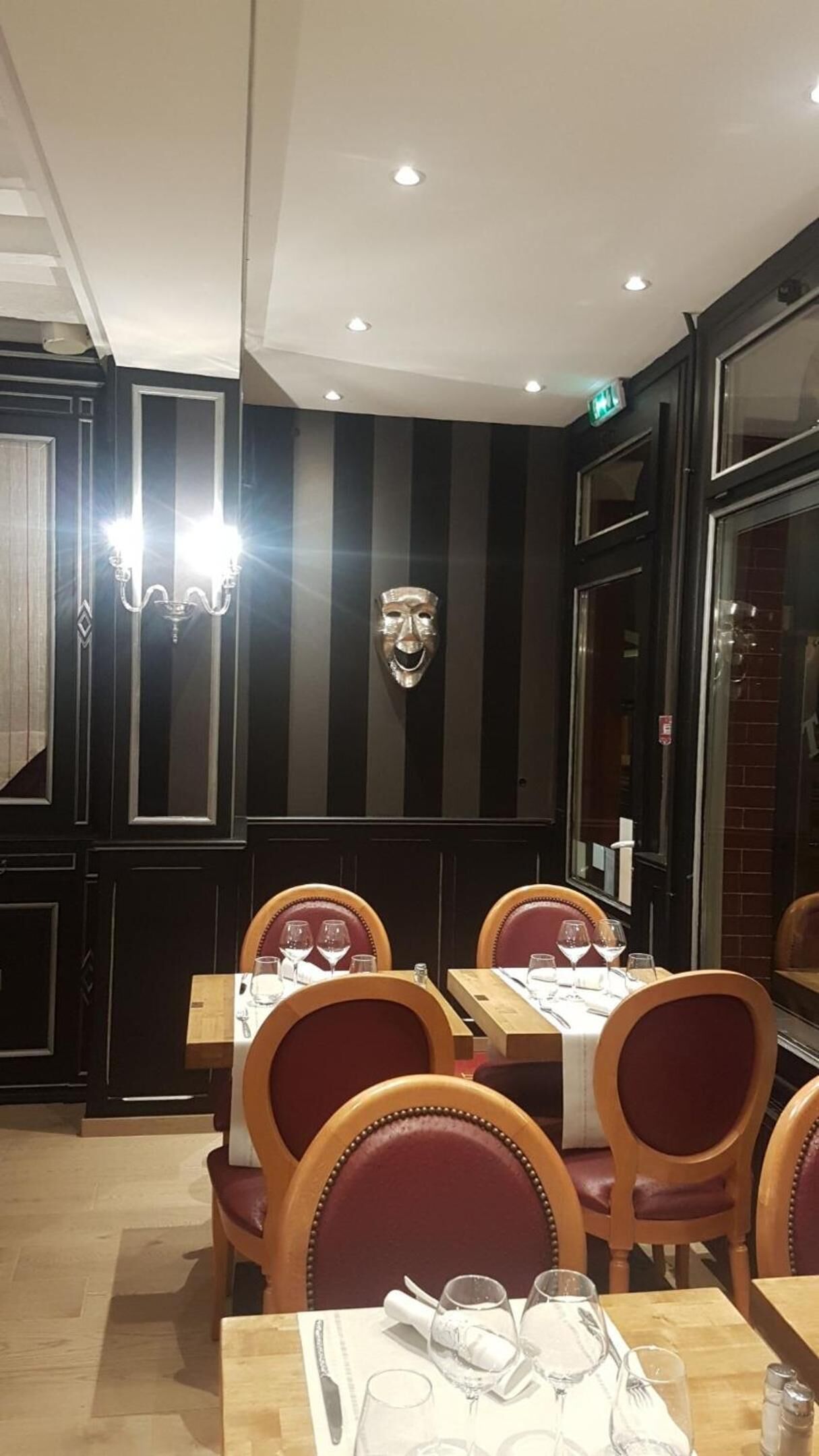 A restaurant interior with dark wood paneling, striped wall decor, and a central theatrical mask. Tables are set with wine glasses and cutlery.