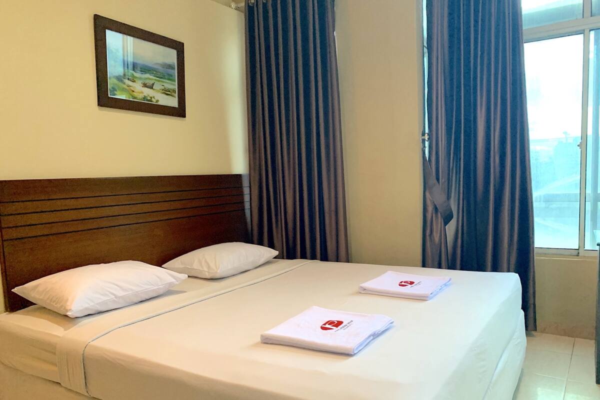A neatly made bed with white linens and pillows, a framed picture on the wall, and a window with curtains.