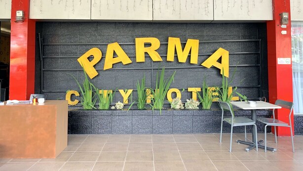 Una mesa de recepción, un cartel con 'PARMA CITYHOTEL' en letras amarillas grandes, y un área de estar con una mesa y sillas.