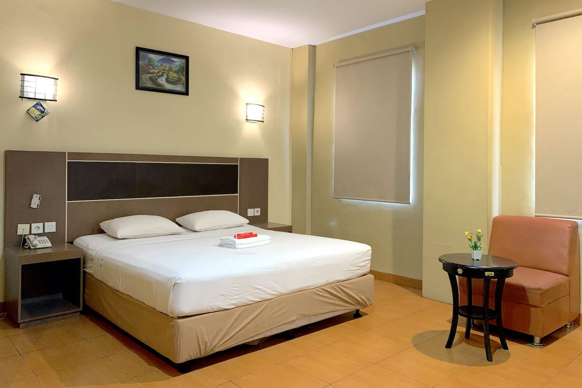 A hotel room with a large bed, bedside tables, a small table with a chair, a window with a blind, and a painting on the wall.
