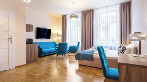 A modern hotel room with a wooden herringbone floor, a blue sofa, a pink sofa, and two blue armchairs. There is a flat-screen TV mounted on the wall, a desk with a lamp, and a large window with curtains. - Kosmopolita Apartments (Kraków)