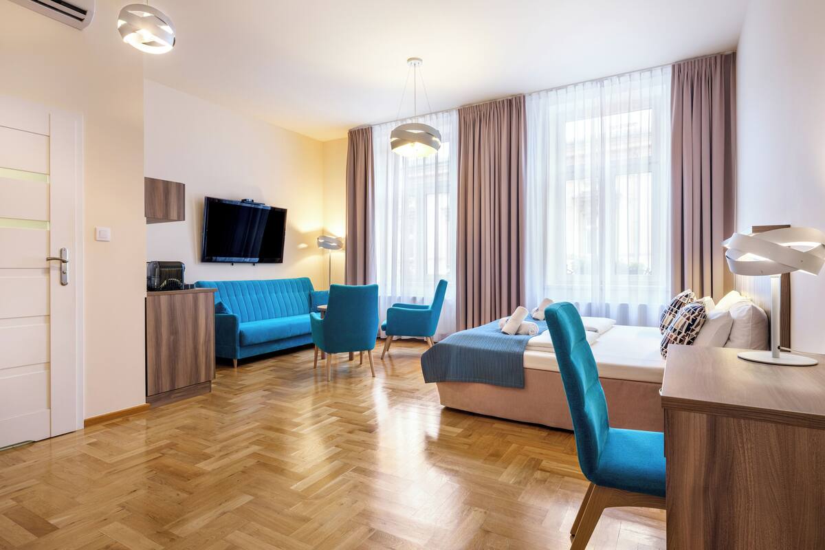 Una habitación de hotel moderna con piso de madera en espiga, un sofá azul, un sofá rosa y dos sillones azules. Hay un televisor de pantalla plana montado en la pared, un escritorio con lámpara y un ventanal con cortinas.