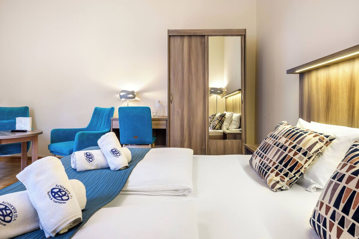 Habitación de hotel con una cama, dos sillas azules, un escritorio, un espejo y toallas con un logotipo.