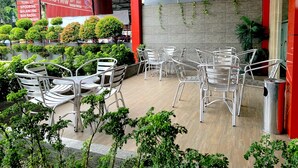 Un espace extérieur aménagé avec des chaises et des tables en métal, entouré de verdure et bordé d’un mur rouge.