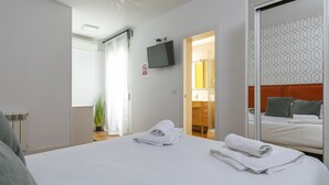 Habitación de hotel con una cama, un televisor montado en la pared, un baño a la vista a través de una puerta abierta y una planta cerca de la ventana.