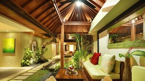 A spacious living area with a high ceiling, a wooden ceiling, a sofa with cushions, a coffee table with a floral centerpiece, and a view into the adjacent room. - The Hotman Paris II Seminyak (Seminyak)