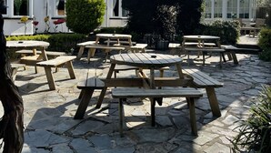 Een binnenplaats met stenen vloer en houten picknicktafels, omgeven door een wit gebouw met een balkon en een conservatorium.