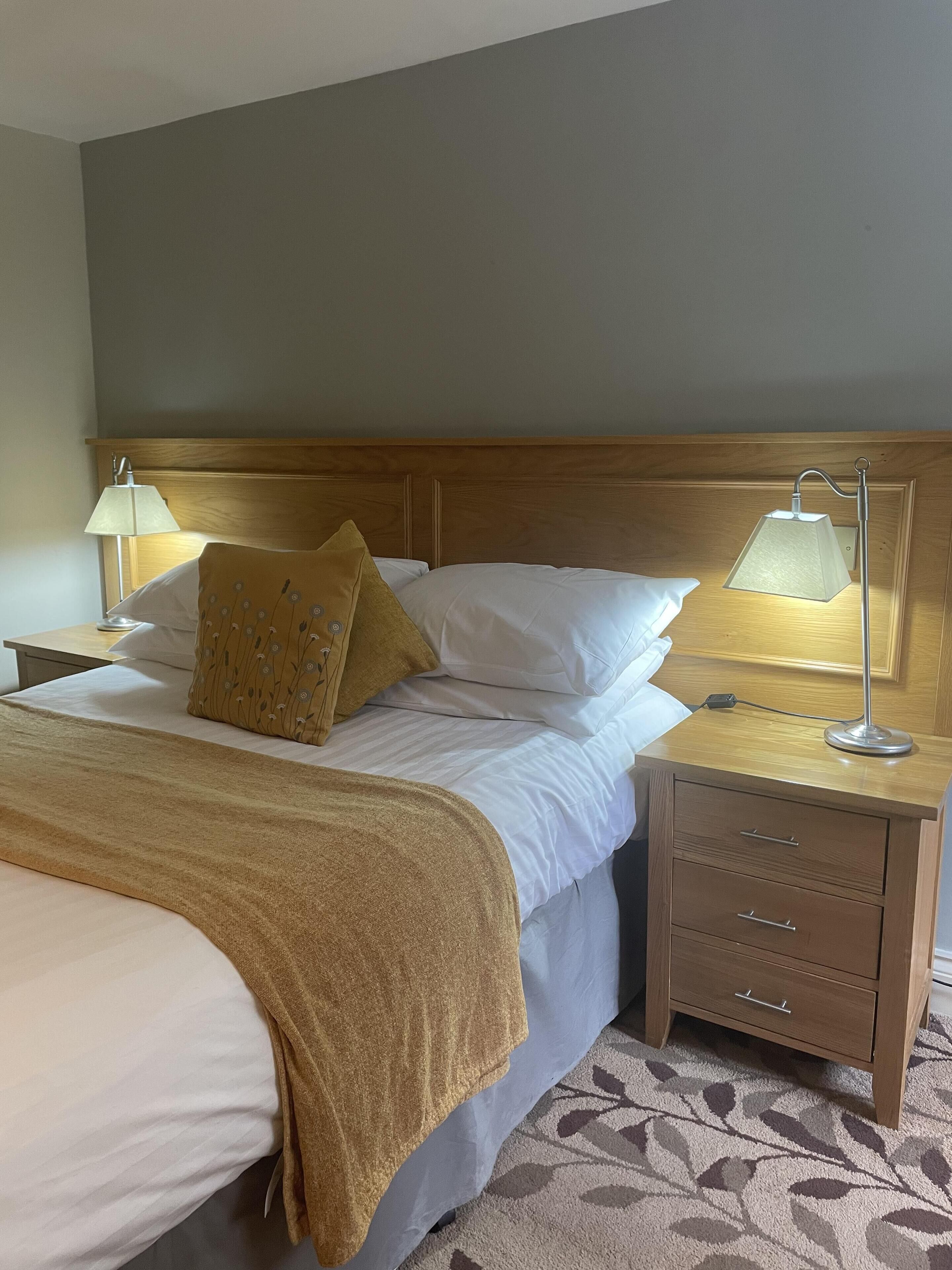 A hotel room with a bed, two bedside tables, a lamp on each table, a wooden headboard, and a patterned carpet.