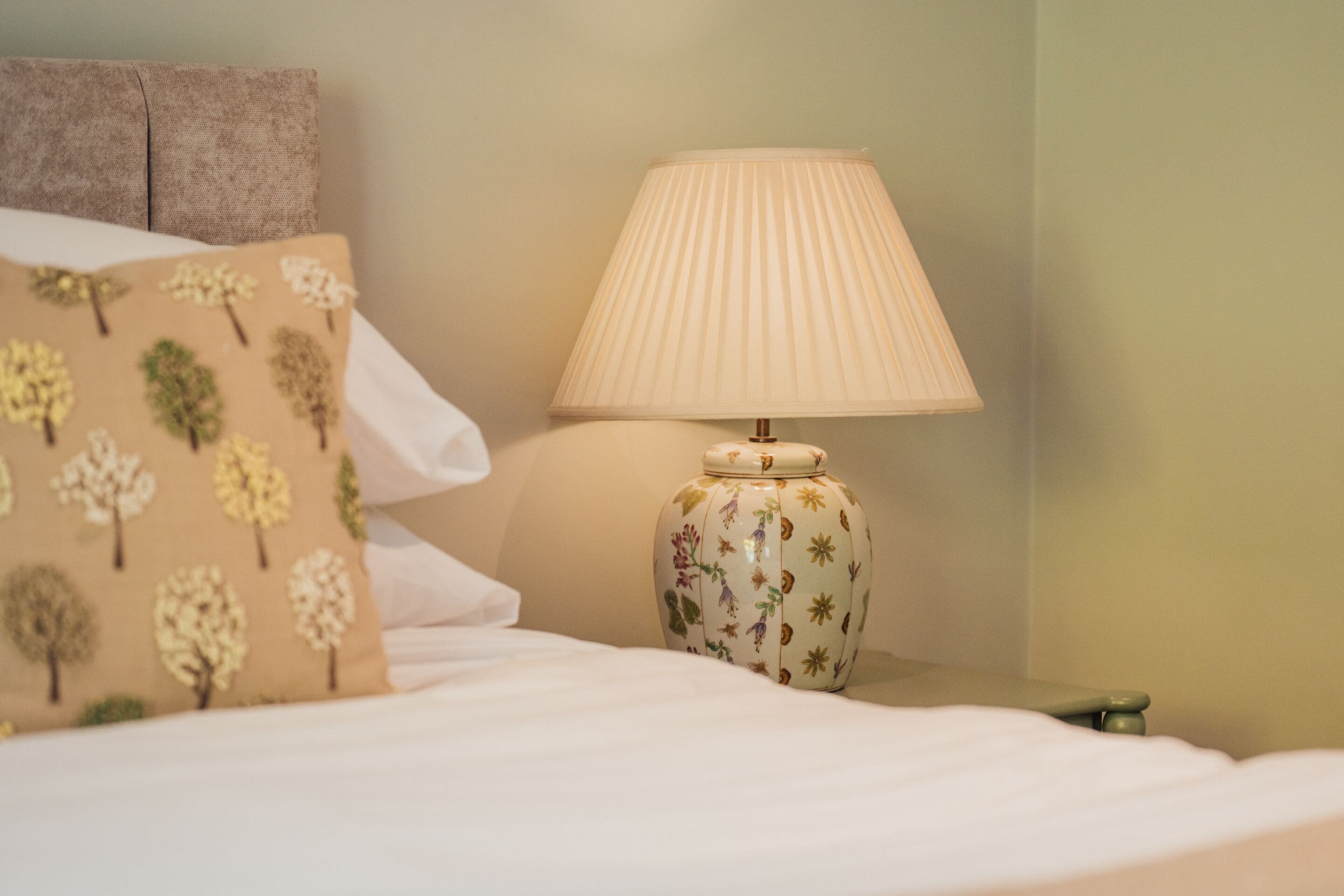 Un lit avec un oreiller blanc et un oreiller beige orné de motifs d’arbres, une lampe blanche avec un abat-jour plissé et une table de chevet verte.